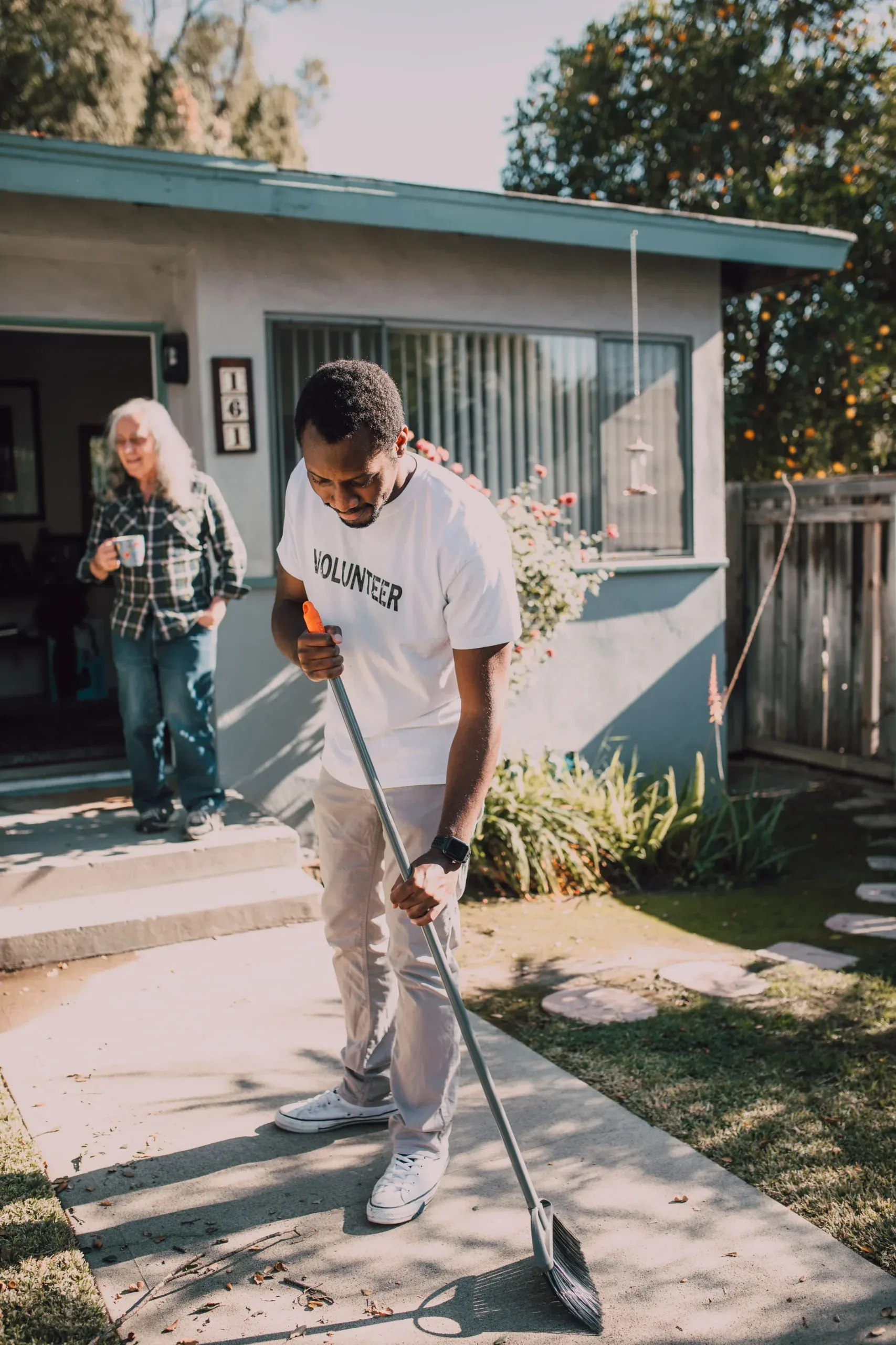A volunteer sweeps a garden path outside a home while a woman holds a mug nearby.