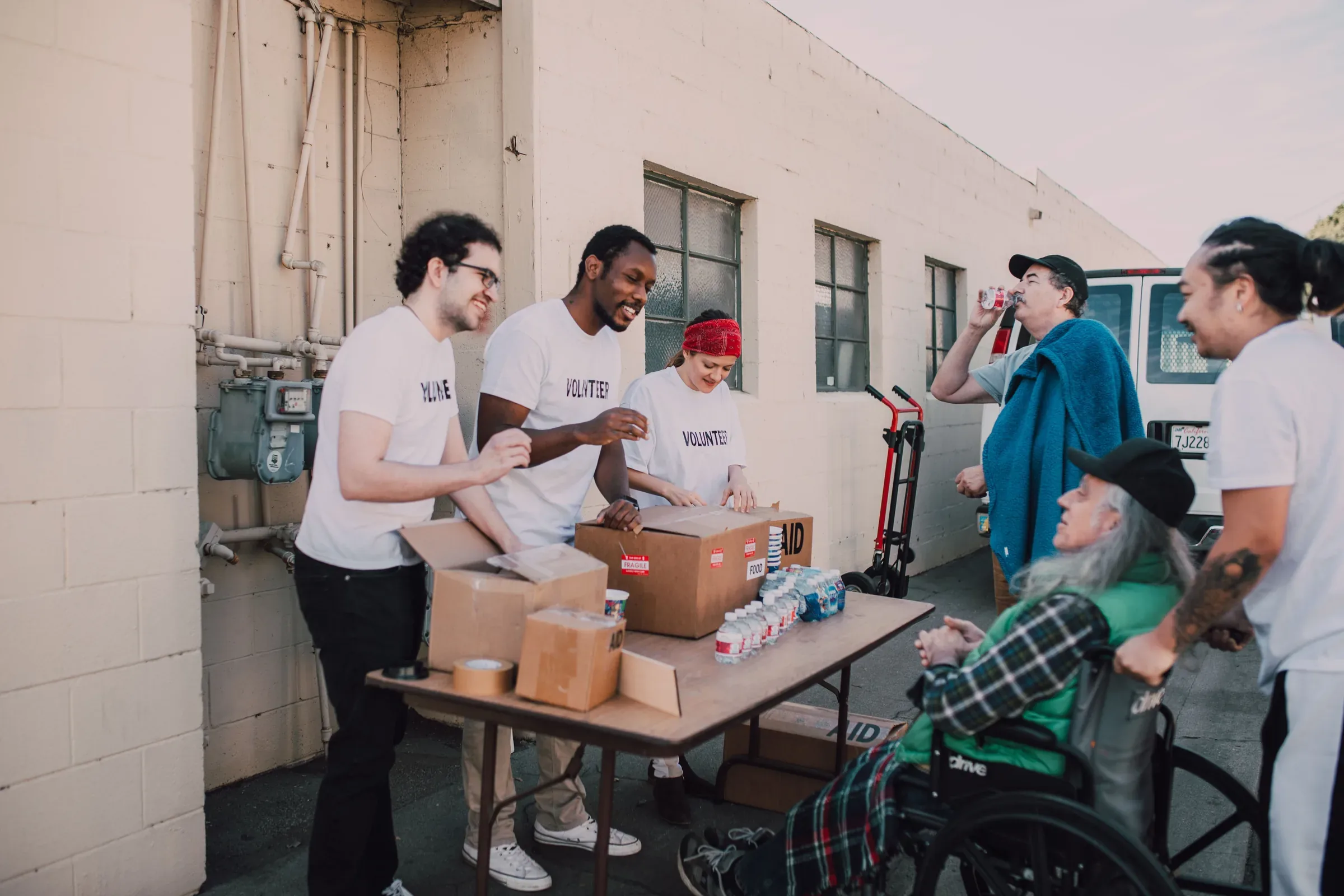 Volunteers in white shirts pack food boxes beside older adults in an outdoor alley