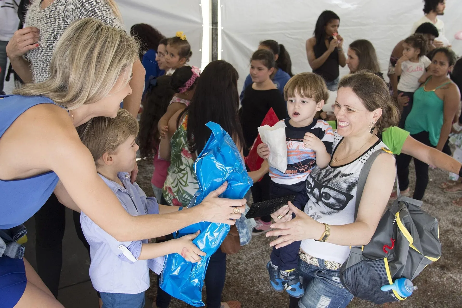A woman hands a blue gift bag to a boy while another woman holds a toddler in a crowd.
